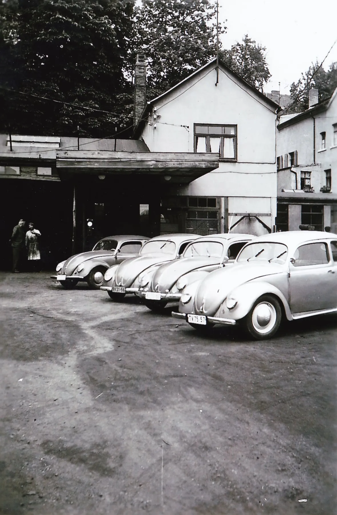 Reihe geparkter VW Käfer auf dem Hof vor dem Wohn- und Werkstattgebäude